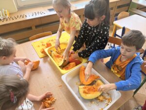Na fotografii widać piątkę dzieci, które znajdują się przy stoliku. Każde z dzieci wybiera pestki z okrojonej na ćwiartki dyni