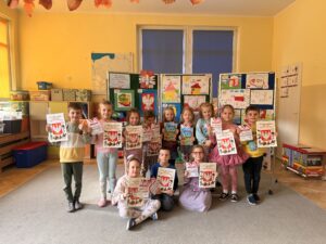 Na fotografii widać grupkę dzieci, która prezentuje książki/kolorowanki o Polsce i dyplomy za udział w konkursie plastycznym. W tle tablice z pracami plastycznymi.