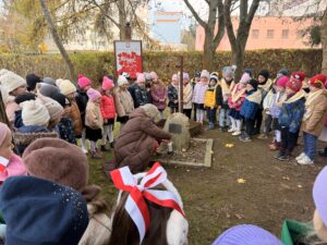 Na fotografii widać dzieci w zimowych ubraniach, zgromadzone wokół dębu katyńskiego i kamiennego pomnika. W tle widać przedszkolny ogród i drzewa. W centrum fotografii znajduje się jedna z nauczycielek, zapalająca znicz.