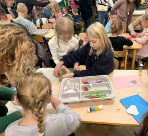 Na fotografii widać dzieci z rodzicami, siedzących przy stolikach i ozdabiających kolorowe kartki bożonarodzeniowe. Dzieci wraz z rodzicami korzystają z klejów, nożyczek i brokatowych ozdób, przyklejając je na kartkach.
