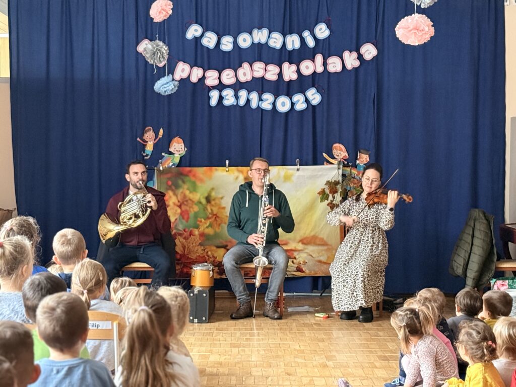 Na fotografii widać dzieci siedzące w sali gimnastycznej tyłem do trzech muzyków, którzy siedzą na tle granatowej kotary. Muzycy grają na waltorni, klarnecie basowym i skrzypcach.