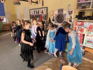 Na zdjęciu widać dziewczynki, przebrane za Elsa z krainy lodu, czarodziejki i inne magiczne postacie. Dziewczynki są ustawione w rzędzie, obok rzędu stoją buty dzieci, również ustawione w kolejce. Dziewczynki biorą udział we wróżbie "wyścig kapci".