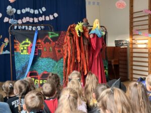 Na zdjęciu widać dzieci siedzące tyłem do fotografa. W tle dwie aktorki w czerwonych sukniach. Aktorki trzymają kukiełkę smoka i kukiełkę księżniczki. Aktorki stoją na tle malowidła przedstawiającego zamek wawelski, wzgórza, domy i smoczą jamę