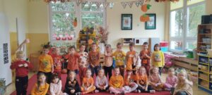 Opis zdjęcia: Grupa dzieci w znacznej większości ubrana w kolorze pomarańczowym na tle kącika przyrodniczego. Chłopcy stoją, dziewczynki siedzą przed nimi.