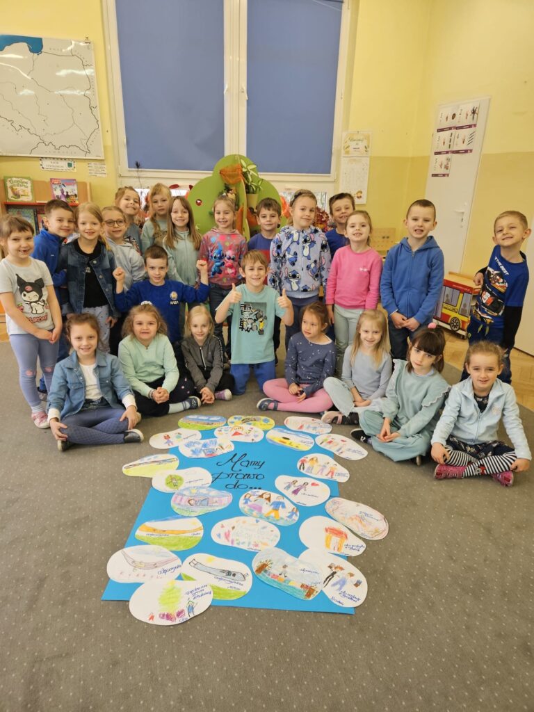 Na fotografii widać dzieci siedzące w gromadce. Przedszkolaki ubrane są na niebiesko, uśmiechają się do fotografa. Na dywanie przed dziećmi leży plakat na którym zobrazowane zostały wybrane przez dzieci prawa dziecka, m.in. prawo do odpoczynku, tajemnicy, spędzania czasu z rodziną.