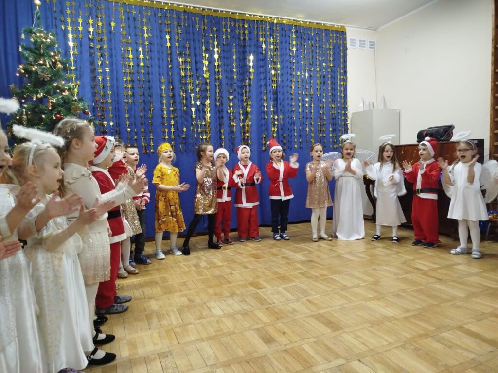 Na zdjęciu widać dzieci stojące w półkolu. Przedszkolaki przebrane są za Mikołajów, aniołki i gwiazdki. Dzieci stoją w sali gimnastycznej, w tle widać granatową kotarę przyozdobioną złotymi gwiazdami.