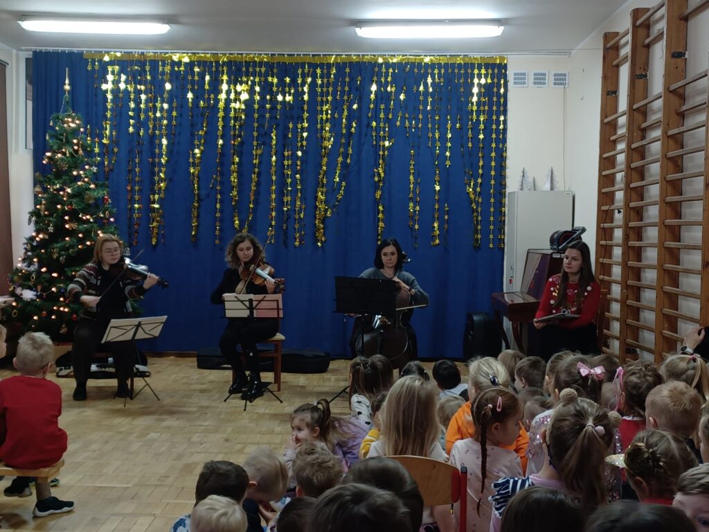 Zdjęcie przedstawia trzech Muzyków grających na skrzypcach, altówce i wiolonczeli. Po prawej stronie sceny muzyki słucha prowadząca koncert. Muzycy usytuowani są na tle świątecznej dekoracji - po lewej stronie choinki oraz złotych gwiazdek na granatowym tle. Widoczne są również dzieci podziwiające występ.