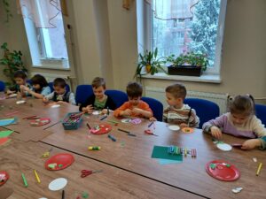 Dzieci siedzącą przy stołach i tworzą bombki poprzez ozdabianie papierowych i piankowych szablonów. Na stołach rozłożone są przybory plastyczne (żele brokatowe, kartki, nożyczki, naklejki, itp.), z których dzieci swobodnie korzystają.