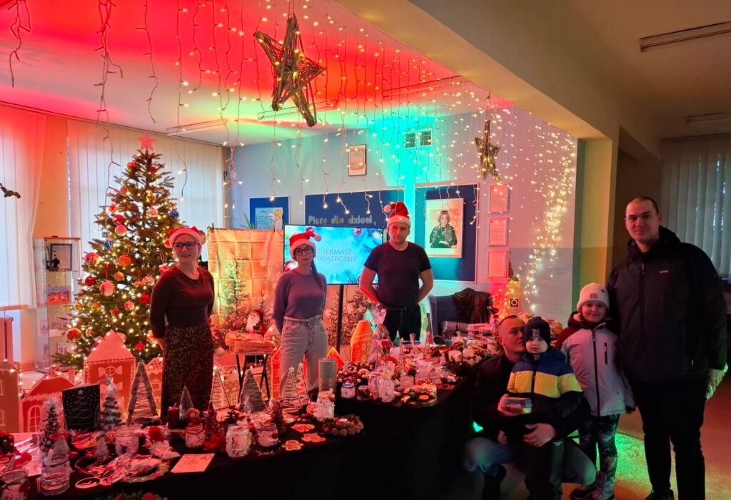 Zdjęcie przedstawia udekorowany hol przedszkola - pełen światełek, w którym ustawione jest stoisko z ozdobami Bożonarodzeniowymi. Stoiska dogląda troje rodziców w mikołajowych czapkach, natomiast dwoje dzieci z rodzicami zatrzymało się przy wystawionych dekoracjach.