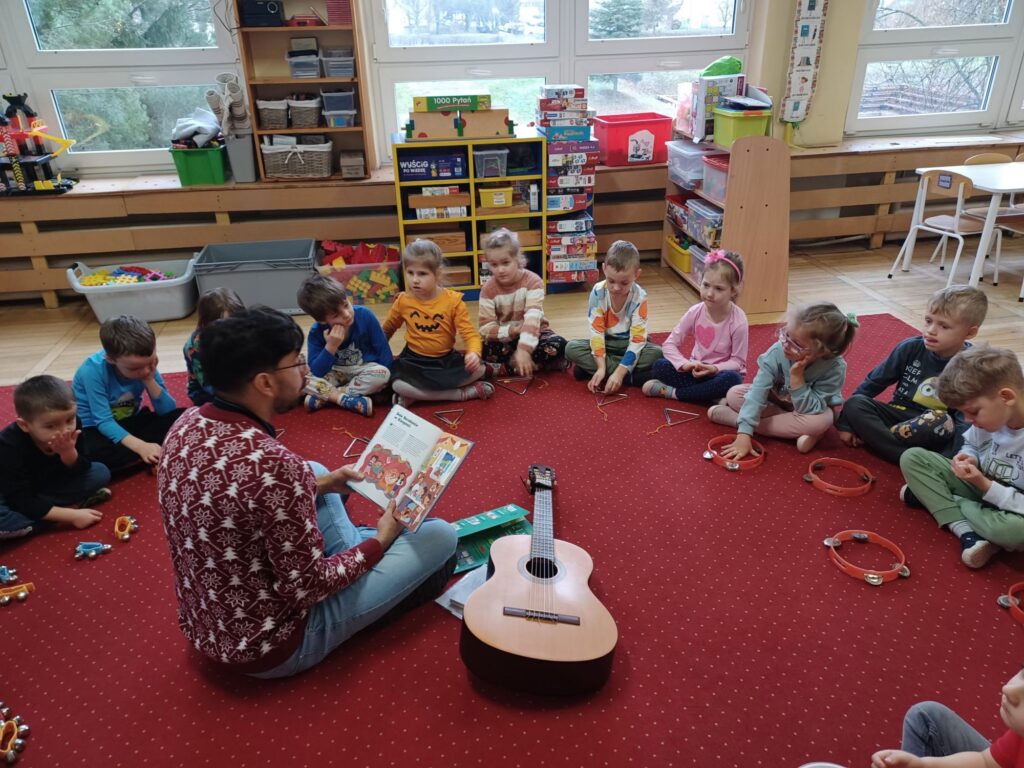 Zdjęcie przedstawia dzieci siedzące w kole ma dywanie oraz Tatę Kajetana siedzącego wewnątrz koła i czytającego książkę. Przed dziećmi leżą instrumenty, które wcześniej były wykorzystywanych do zabaw muzycznych. Obok Gościa - po prawej stronie, leży gitara.