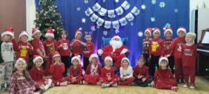Grupa dzieci ubrana w kolorze czerwonym i w mikołajowych czapkach. Po środku siedzi w fotelu Mikołaj. W tle widać choinkę oraz napis Witaj Mikołaju!